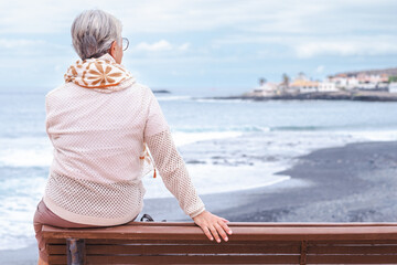 Rear view of senior woman wearing eyeglasses sitting on outdoors bench at sea looking away enjoying freedom and vacation. Active elderly woman relaxing on a sunny day close to the beach