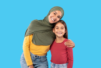 Happy Middle Eastern Mother Wearing Hijab Embracing Daughter, Blue Background