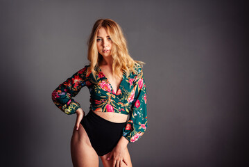 Young blonde woman leaning on one hip in a blouse with a floral print on a gray background