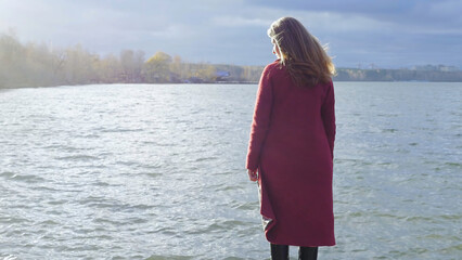 Young girl with her hair stands near the water in the cold season. Back view. Young woman standing...