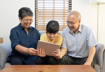 Smiling Asian grandparents on couch with granddaughter looking at tablet. happy three generation family spending time together at home.