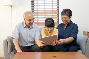 Smiling Asian grandparents on couch with granddaughter looking at tablet. happy three generation family spending time together at home.
