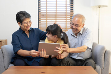 Fototapeta premium Smiling Asian grandparents on couch with granddaughter looking at tablet. happy three generation family spending time together at home.