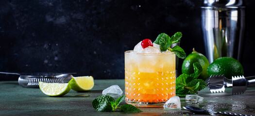 Mai Tai popular alcoholic cocktail with rum, liqueur, syrup, lime juice, mint and crushed ice. Dark green banner background, steel bar tools, negative space