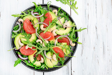 Healthy vegan salad with arugula, avocado, pink grapefruit, cashew nuts and dressing of olive oil, honey and wine vinegar. White rustic kitchen table, top view, negative space