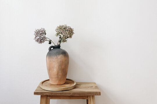 Elegant minimal summer, fall still life photo. Rustic clay vase, pitcher with hydrangea flowers on old wooden stool, console table. White wall background. Empty copy space. Elegant interior, lifestyle