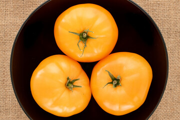 Three organic yellow tomatoes in a black ceramic dish on a jute cloth, close-up, top view.