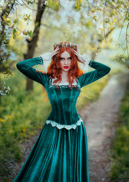 Portrait fantasy red-haired woman medieval queen touches with hands straightens golden crown on head. Girl redhead princess. Green vintage long dress curly hairstyle. Summer nature garden forest trees