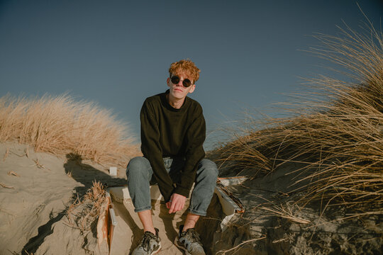 a guy in round glasses and a black jacket sits on the sand dunes