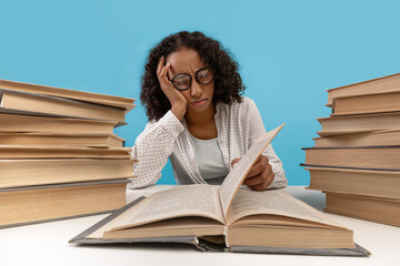 Tired young black female student surrounded by stacks of books reading textbook, getting ready for exam, feeling bored