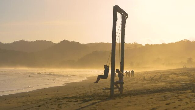 MOVING SHOT: Two friends on big swing on a misty tropical beach at sunset. Playful time on swing on the hazy beach Playa Venao. Carefree leisure activities while travelling through tropical Panama.