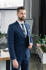 bearded economist looking away while standing with laptop in office.