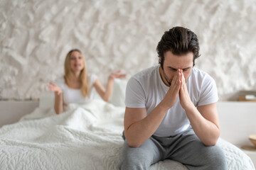 Young man feeling depressed, sitting on bed at home, his angry wife shouting at him, free space. Family problems concept