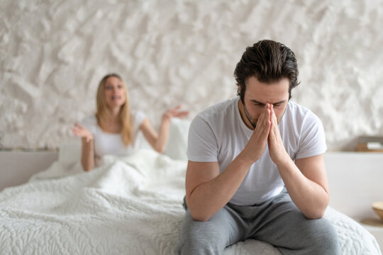 Young Man Feeling Depressed, Sitting On Bed At Home, His Angry Wife Shouting At Him, Free Space. Family Problems Concept