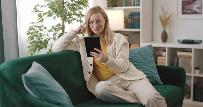 Happy woman in eyeglasses sitting on comfy couch and using digital tablet. Beautiful blonde in casual attire scrolling social media during free time.