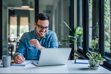 Happy entrepreneur making business plan while working on laptop at home office - obrazy, fototapety, plakaty