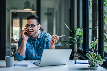 Happy distracted businessman talking on phone while using laptop at home