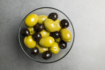 Big green and black olives in glass bowl on stone table, top view.