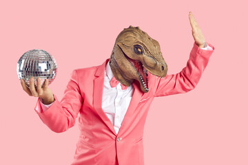 Cheerful, stylish and fashionable man in dinosaur rubber mask with disco ball in his hand on pastel pink background. Man in formal pink suit wears absurd mask on his head and dances. Creative concept.