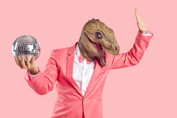 Fotobehang Cheerful, stylish and fashionable man in dinosaur rubber mask with disco ball in his hand on pastel pink background. Man in formal pink suit wears absurd mask on his head and dances. Creative concept. © Studio Romantic