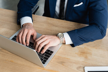 cropped view of businessman in wristwatch typing on laptop in office.