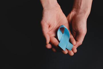 Close up of male hands holding small blue awareness ribbon on black background wall in studio with copy space for promotion content. Symbol of social and medical issues