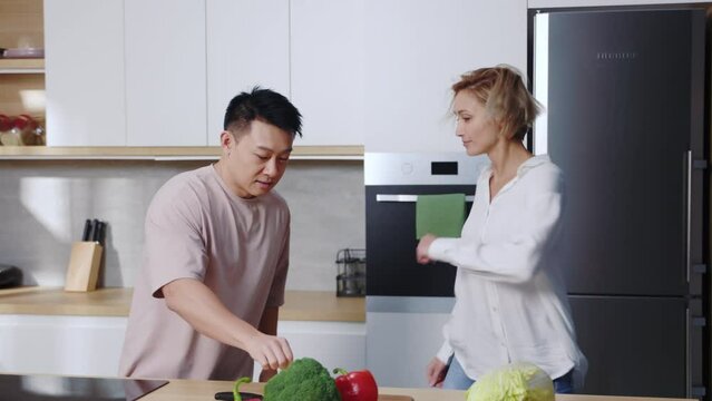 Asian man wearing t-shirt and Caucasian woman with blonde hair dancing in kitchen, high-fiving. Footage of happy multicultural family portrait concept. Excited couple in love. Vegetables on table.
