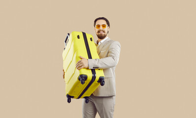 Handsome young businessman going on holiday or business trip. Happy attractive man in formal suit standing isolated on beige background, holding his yellow suitcase and smiling. Travel concept
