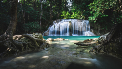 Scenic view of breathtaking waterfall smooth flowing water and emerald green pond in lush rainforest with tree root foreground. Erawan Waterfall, Kanchanaburi, Thailand. Long exposure.