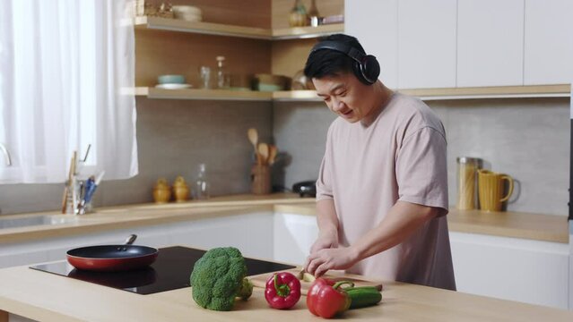 Footage of happy young Asian man in headphones, cutting cucumber. Cute guy wearing casual clothes listening to music while cooking dinner in cozy kitchen of new apartment. Weekend, home. Indoors