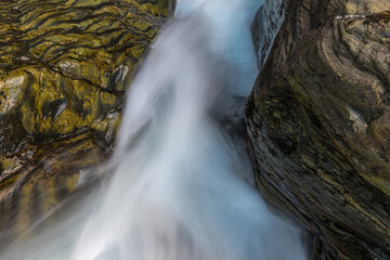 Between rocks falls a raging mountain stream of clear water photographed with motion blur.