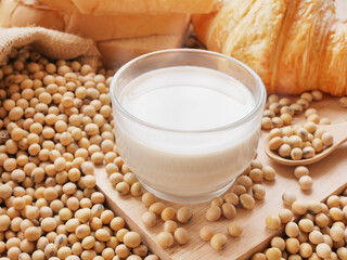Hot cup of soy milk on lots of soy beans on wooden cutting board with sack of soy beans with some croissants and breads