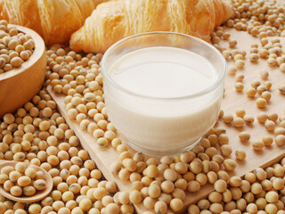 A hot glass soy milk on wooden cutting board with wooden bowl of soy beans and two pieces of croissants
