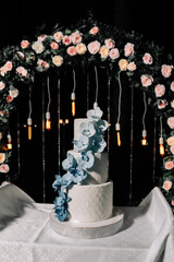 A wedding cake. Sweets. Wedding cake on a black background three floors with white mastic decorated with blue and blue orchids from mastic on the table against the backdrop of the wedding arch