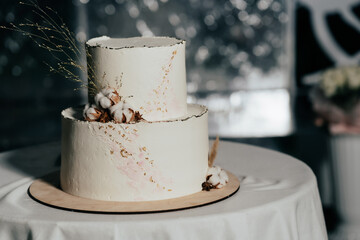 A wedding cake. Milk-colored cake decorated with cotton beads and gold leaf on a dark background in...