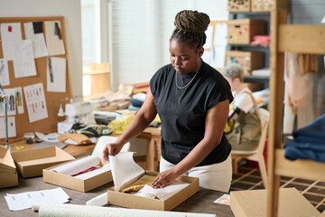 African American woman in casualwear standing by table and packing new clothes in cardboard box...