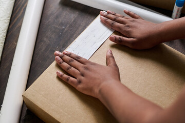 Hands of young African American female putting paper with client address on top of packed box with...