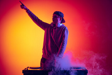 Obraz na płótnie Canvas Portrait of active young man, professional dj playing, making sounds isolated over gradient red yellow background in neon light
