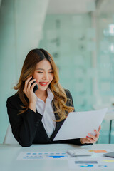 An Asian female accountant sits at a table talking on a smartphone with a laptop and calculates a financial graph showing investment results. Plan a successful business growth process in the office.