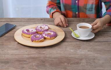 Woman is having happy while drinking coffee, left hand raises white cup of coffee prepare to drink, right hand holds green ceramic spoon with purple donuts placed in  wooden tray next to  mobile phone