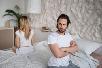 Unhappy young couple having relationship problems, sitting on opposite sides of bed, not looking at...