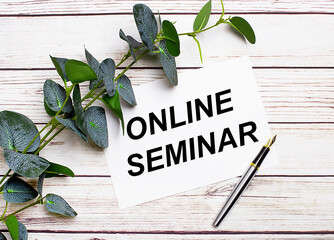 On a light wooden table, there is a eucalyptus branch, a fountain pen and a sheet of paper with the text ONLINE SEMINAR