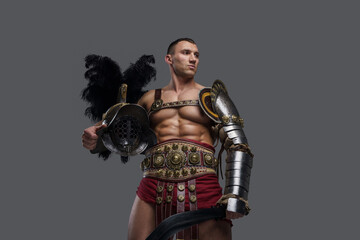 Shot of isolated on grey background gladiator with naked torso holding sword and helmet.
