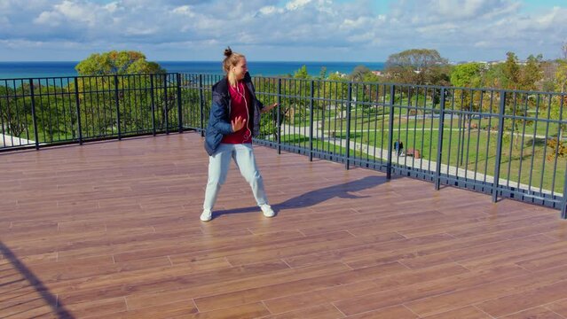 Sportive girl performs modern dances on stage fenced with railing surrounded by trees. Female dancer dancing contemporary, hip hop, shuffle in park. Autumnal sunny day. Creative expression, hobby