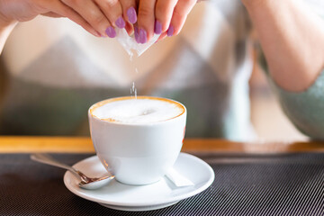 blurred female hands add sugar from a sachet to a white cup of cappuccino in a cafe at the table