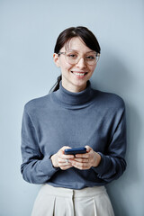 Fototapeta premium Candid portrait of young businesswoman holding smartphone and smiling at camera against simple blue background