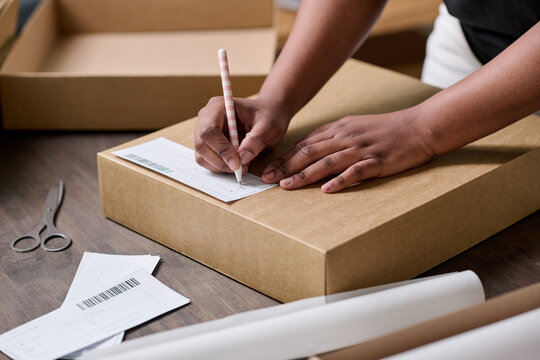 Hands Of Young Black Woman With Pen Writing Down Address Of Client On Top Of Cardboard Box Cover Before Packing Ordered Goods