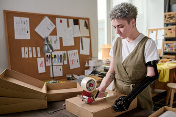 Young serious female worker of warehouse sealing packed box with ordered goods with cellotape...