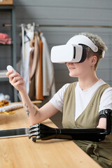 Young modern woman with myoelectric arm in vr headset using remote control while making or...