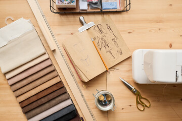 Above angle of wooden table with fabric samples, notebook with sketches and pencil, electric sewing machine, ruler and scissors